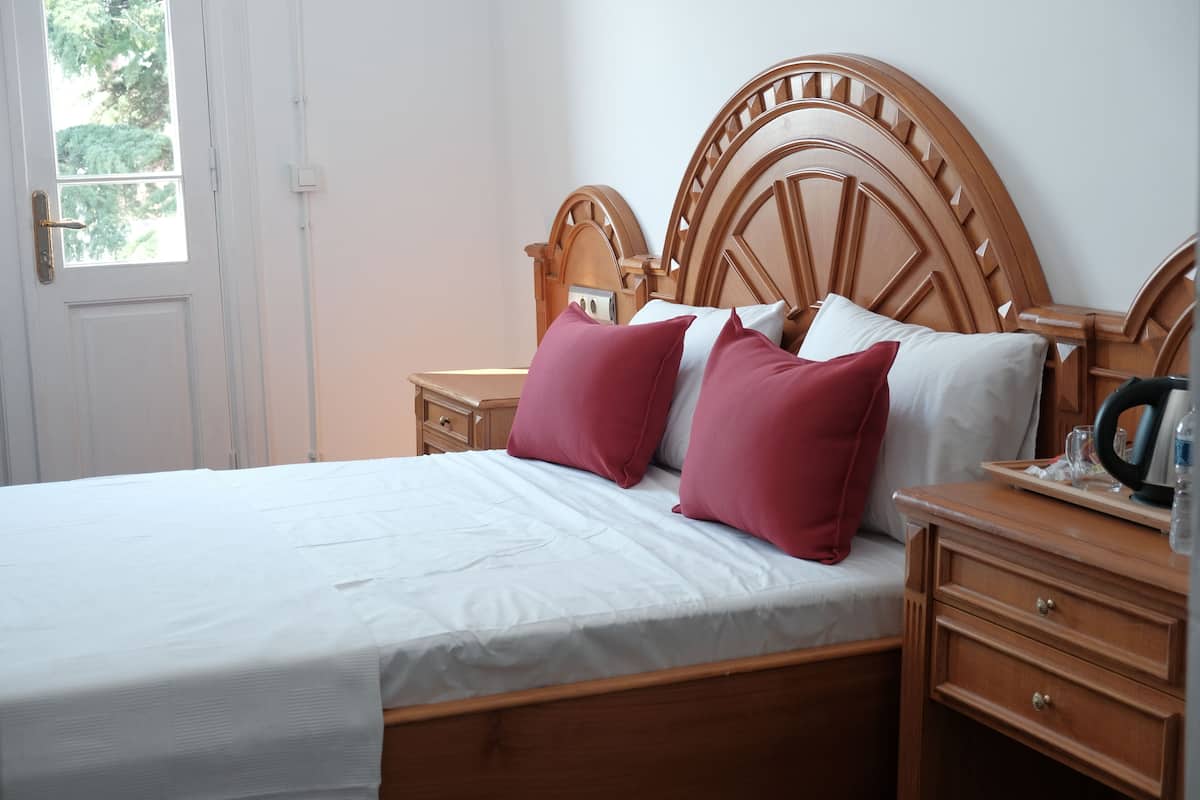 A neatly made bed with a wooden headboard, a bedside table with a tray, and a window with a view of trees.