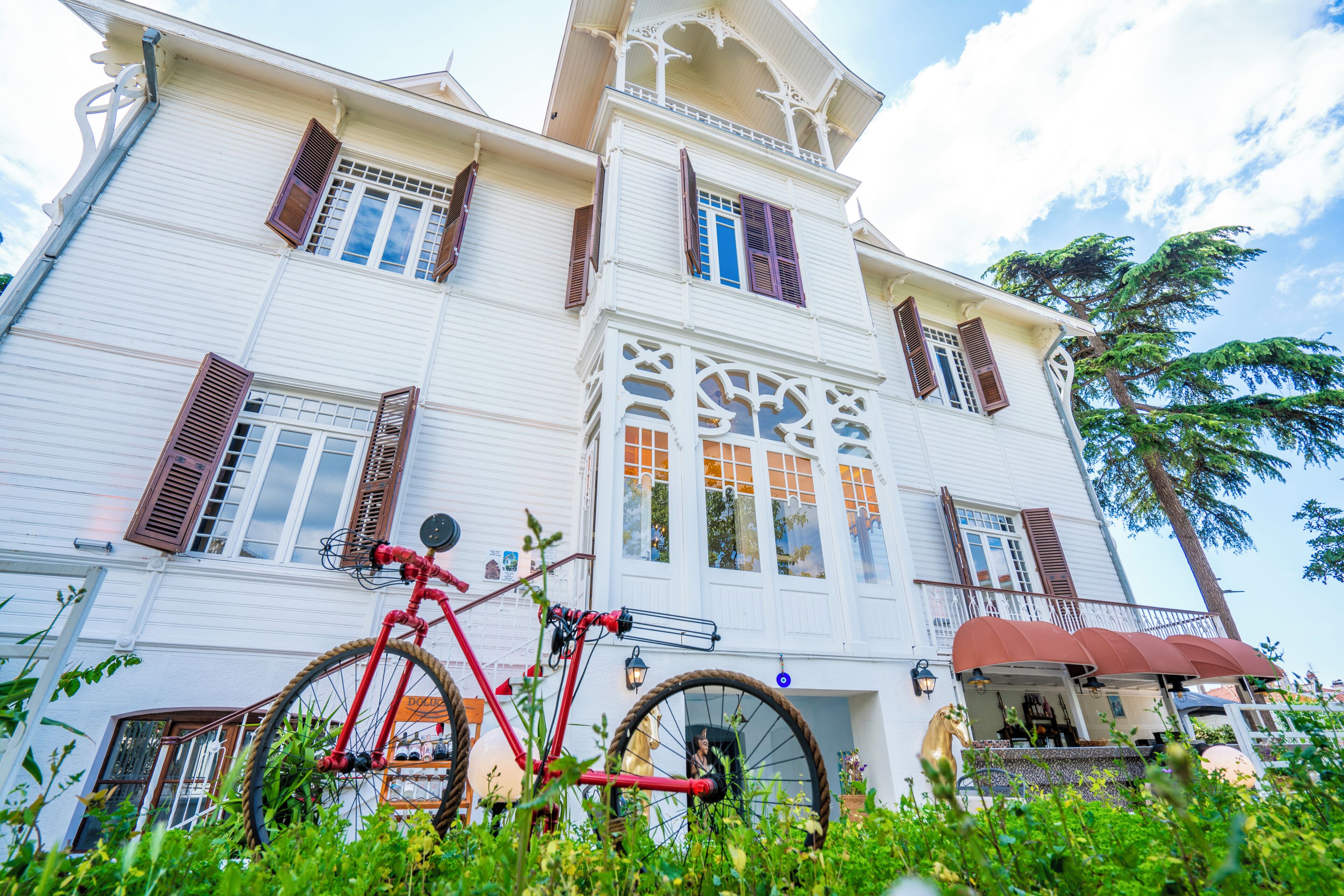 A white building with brown shutters, a red bicycle, and lush greenery in front.