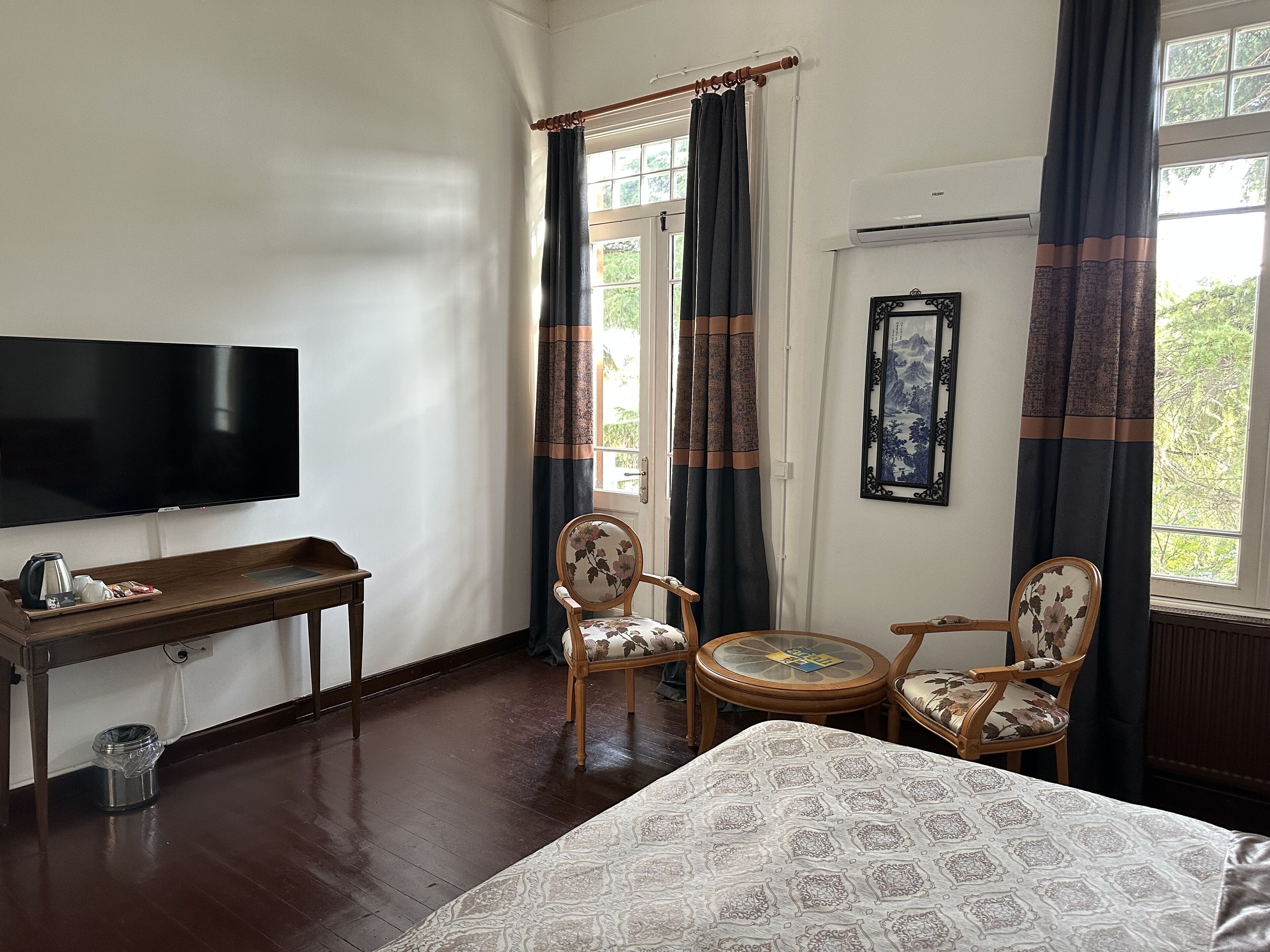 A hotel room with a bed, a TV, a desk with a coffee maker, two chairs, a small table, and a window with curtains.