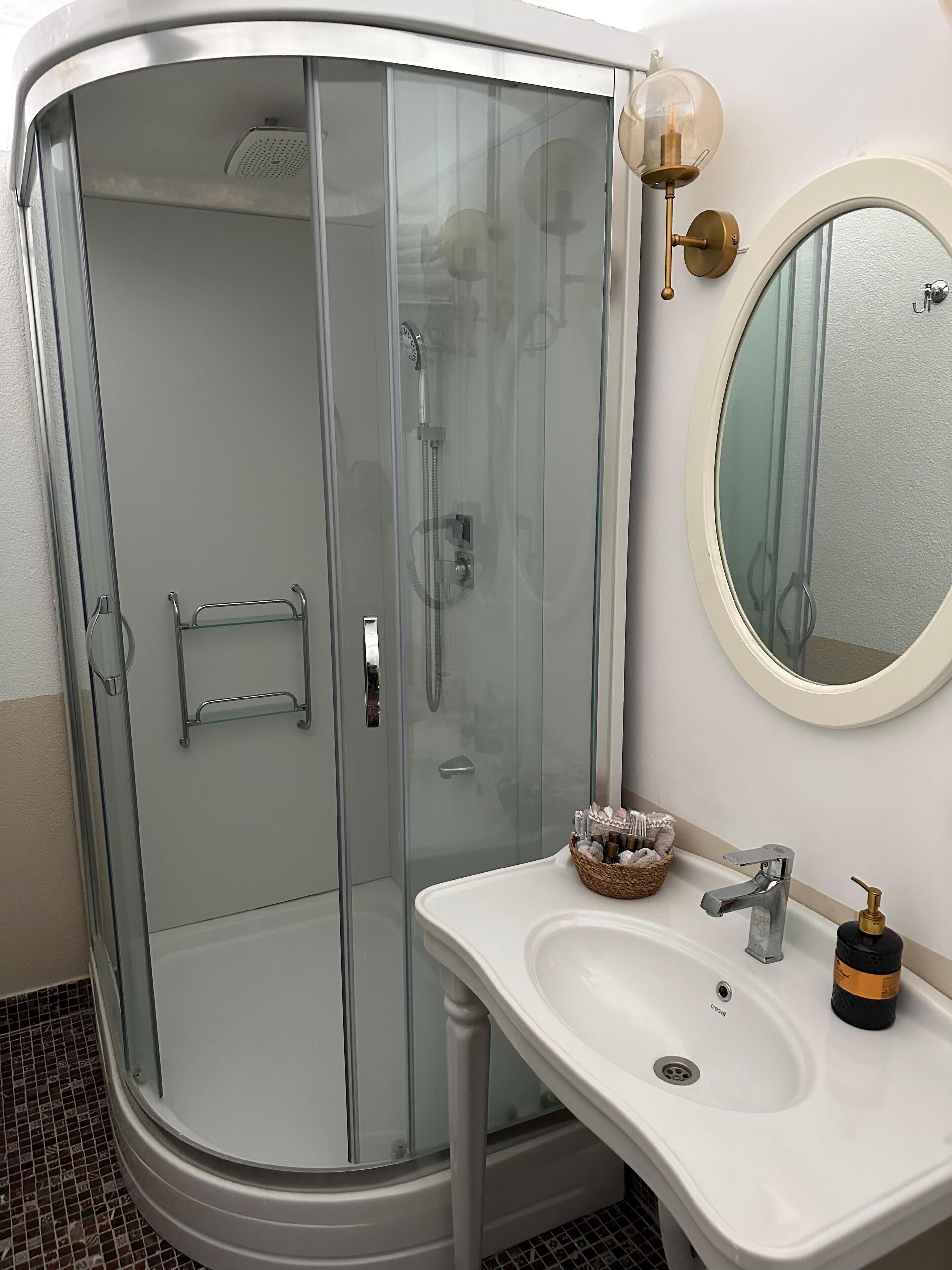 Ein modernes Badezimmer mit einer gläsernen Duschkabine, einem weißen Waschbecken mit goldener Armatur und einem runden Spiegel.