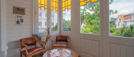 Una habitación con dos sillas de madera y una mesa de vidrio, una puerta blanca con vidrieras amarillas, y vistas a edificios y vegetación.