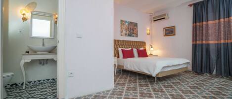 A bedroom with a bed, a bedside table, a nightstand, a wall-mounted lamp, a painting, and a window with curtains.