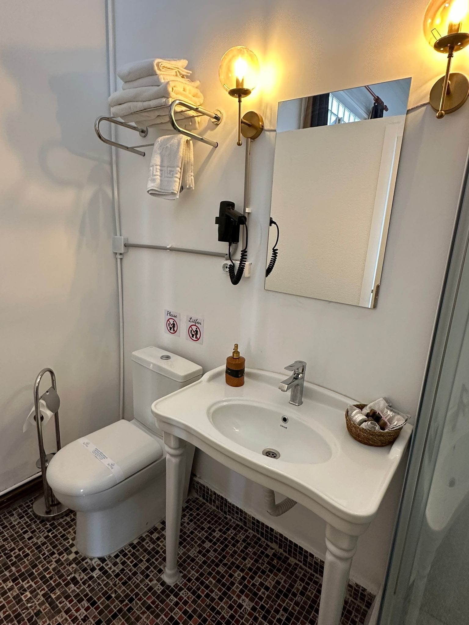 Ein modernes Badezimmer mit Toilette, Waschbecken und Spiegel. Handtücher an einem Ablagegestell, eine Seifenspender und ein Korb mit Pflegeprodukten.
