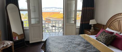 Habitación de hotel con cama, una silla, una mesita de noche, una lámpara y un balcón con vistas a la ciudad.