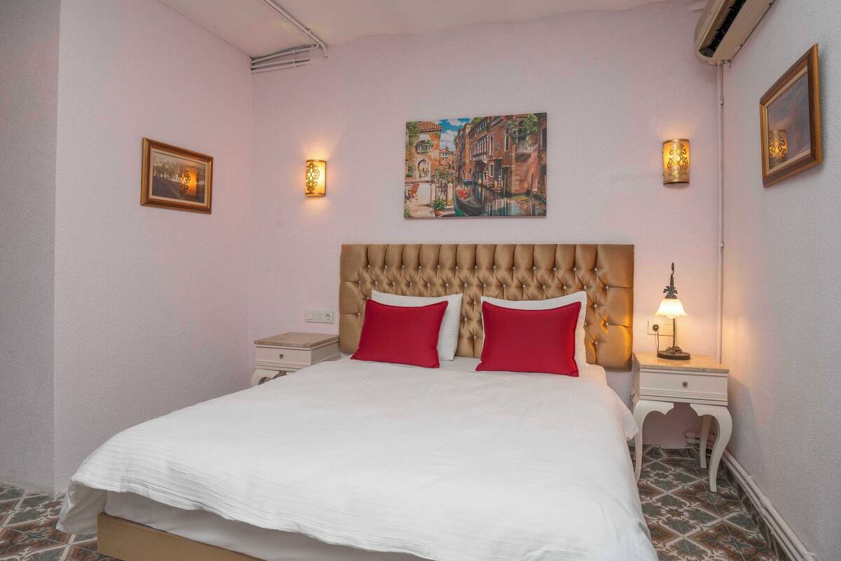 A neatly made bed with a tufted headboard, two red pillows, and a white comforter. There are two bedside tables with lamps, a wall-mounted painting, and a framed picture.