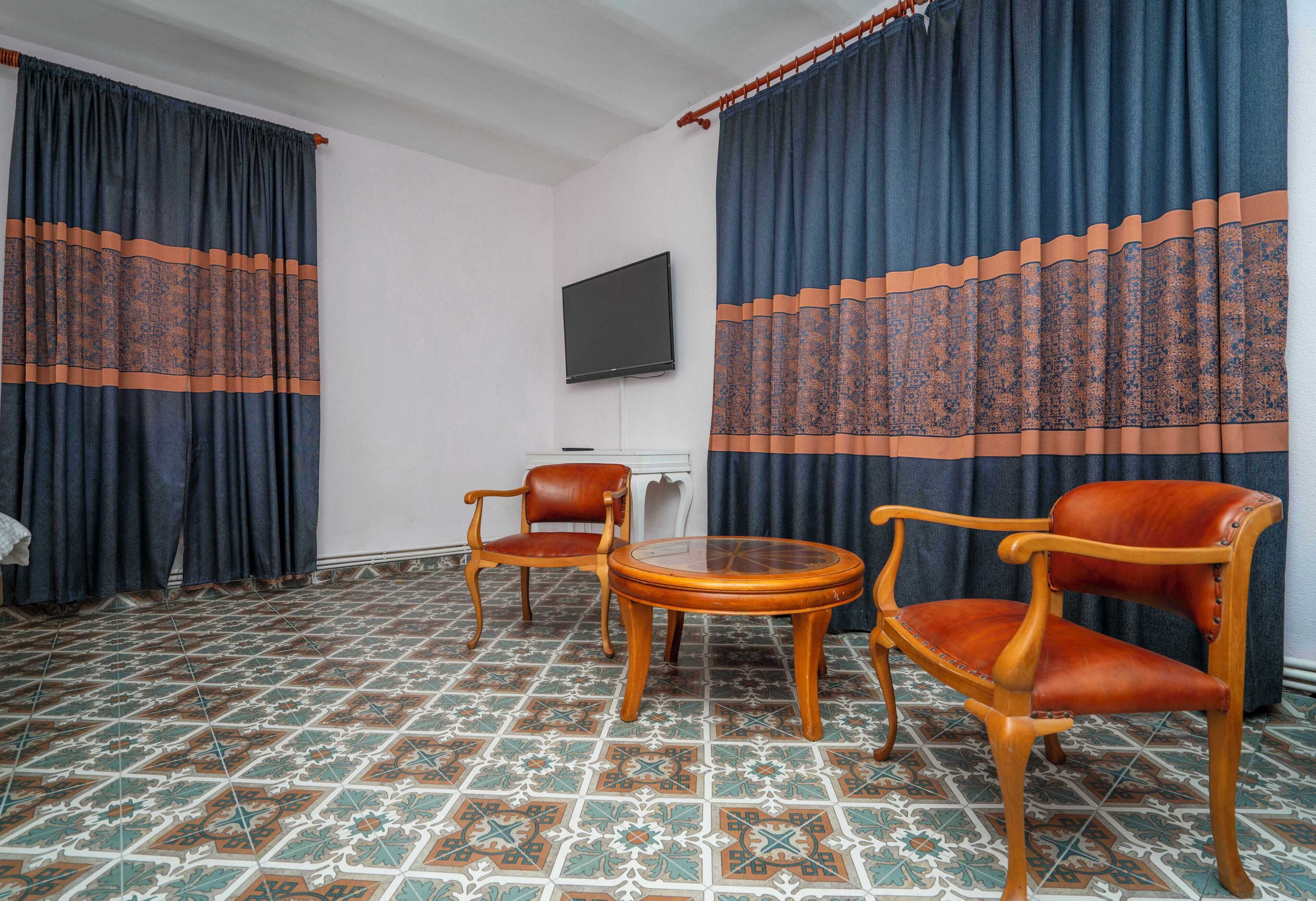 Quarto com duas cadeiras de madeira, uma mesa redonda pequena, televisão de tela plana na parede e piso com azulejos estampados.