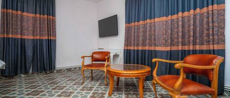 Quarto com duas cadeiras de madeira, uma mesa redonda pequena, televisão de tela plana na parede e piso com azulejos estampados.