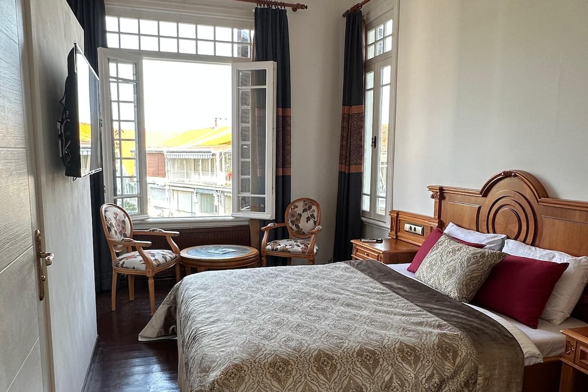 A hotel room with a large bed, a wooden nightstand, a chair, a small table, and a TV mounted on the wall.