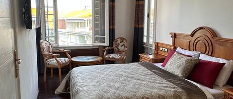 A hotel room with a large bed, a wooden nightstand, a chair, a small table, and a TV mounted on the wall.
