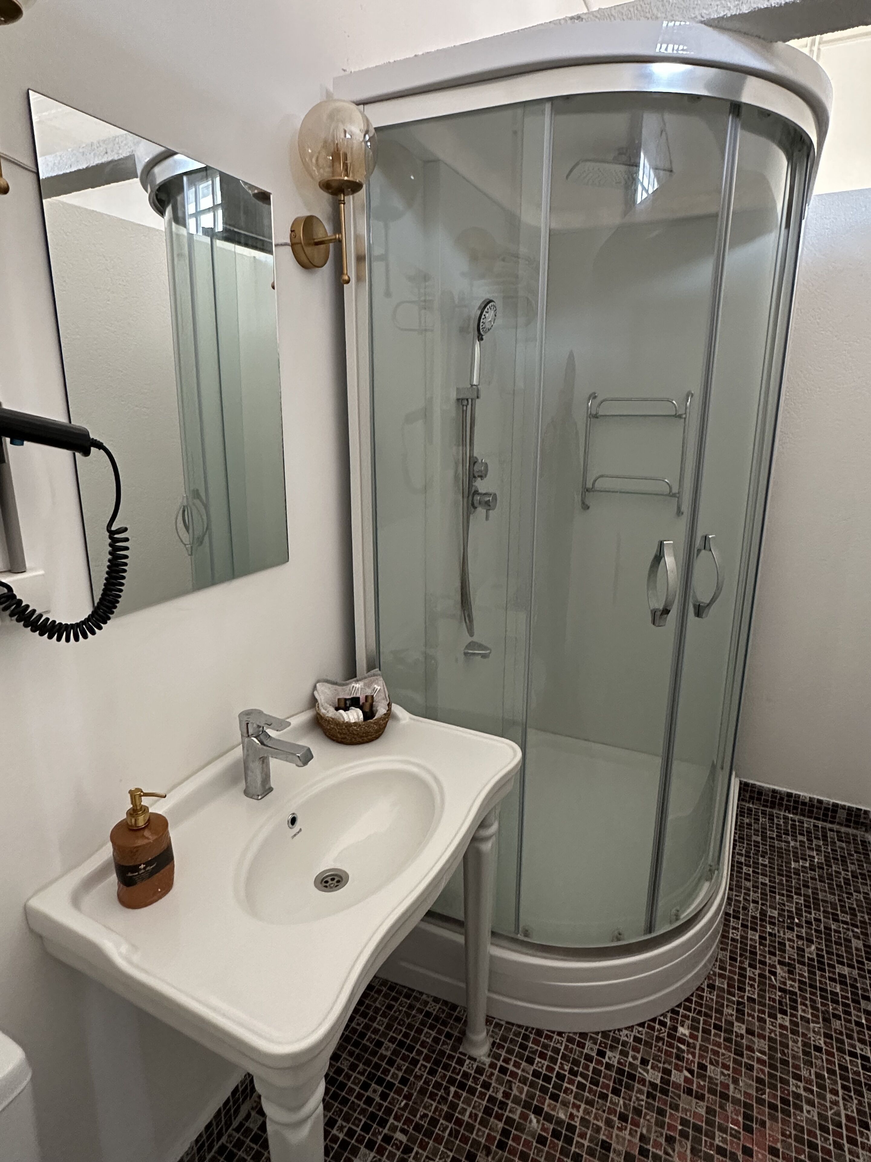 A bathroom with a glass-enclosed shower, a white sink with a soap dispenser, and a mirror on the wall.