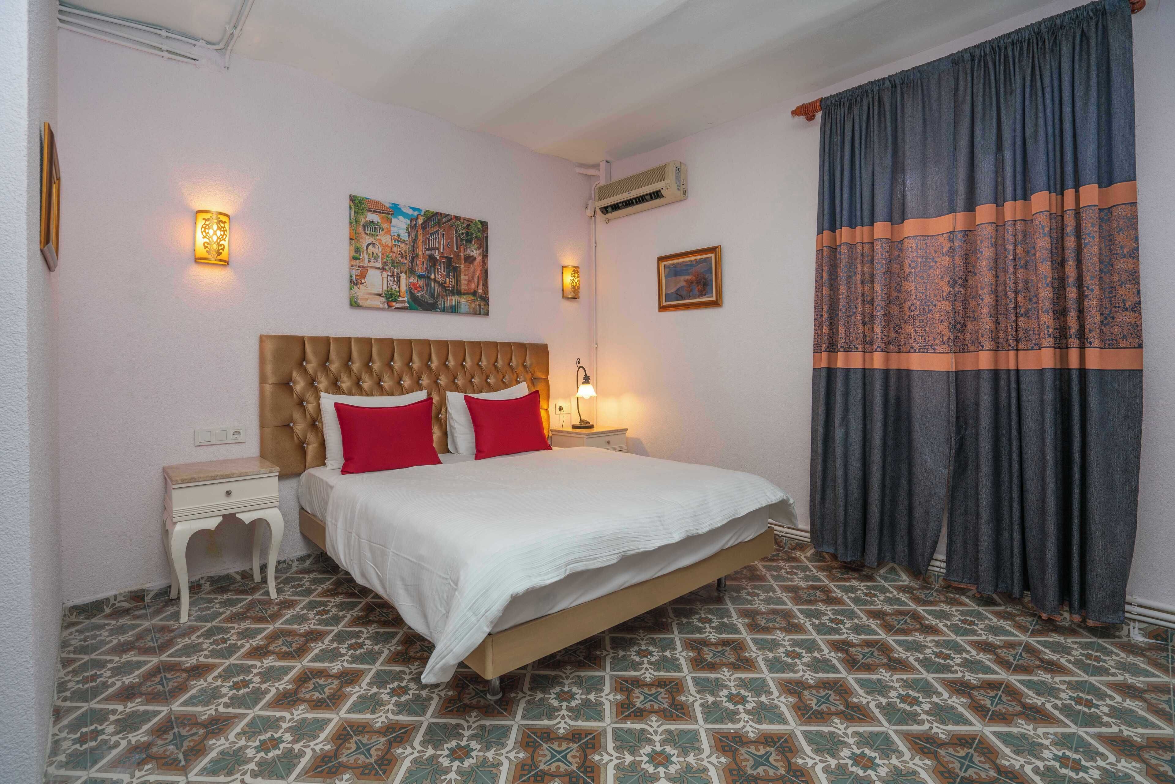 Ein Schlafzimmer mit einer gebügelten Kopfstütze, einem Bett mit weißer Bettwäsche und roten Kissen, einem Nachttisch mit Lampe, gemusterten Bodenfliesen und einem großen Fenster mit Vorhang.