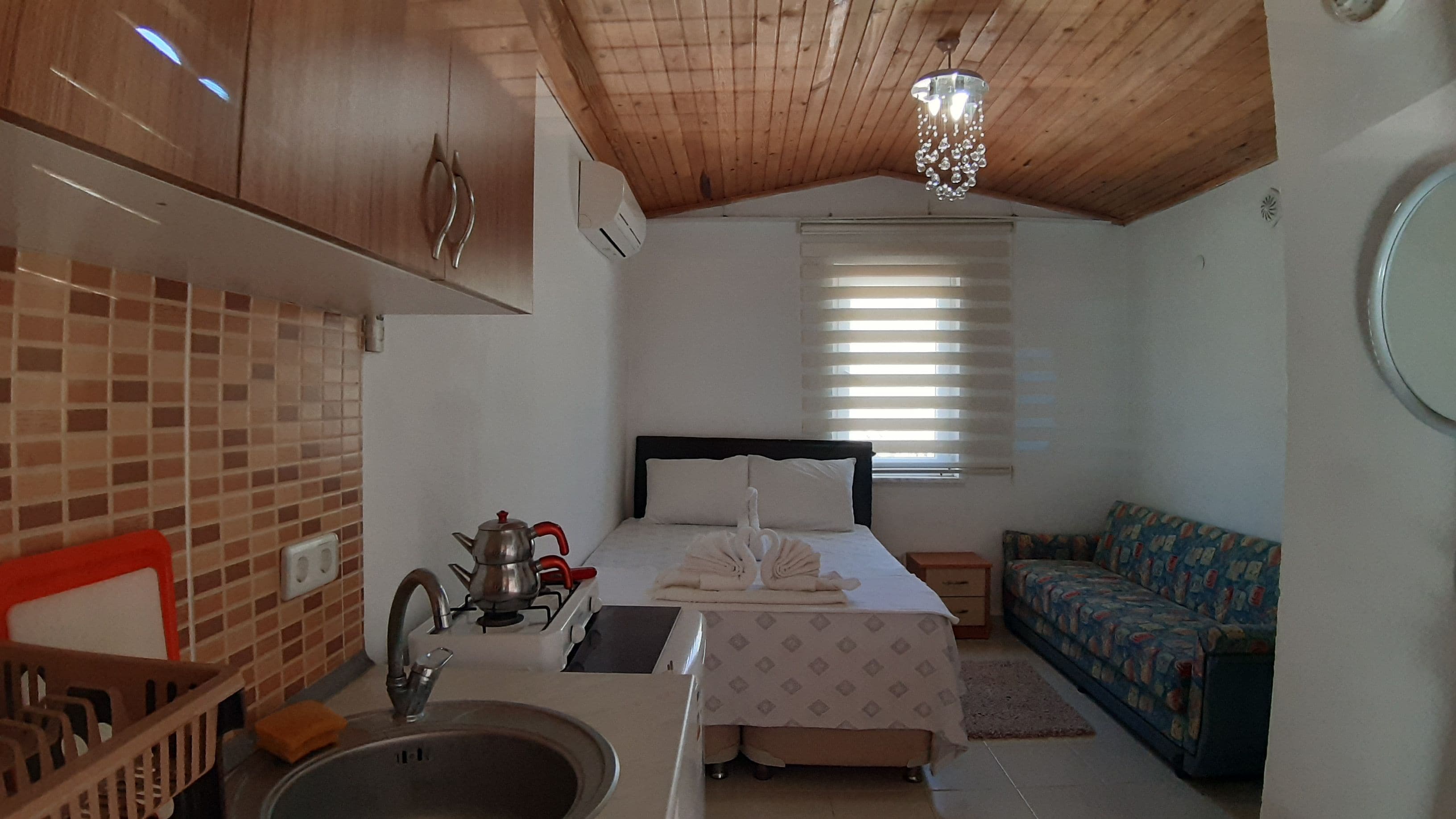 Una habitación compacta con kitchenette, una cama con cubrecama blanco, un sofá estampado y una ventana con persianas.