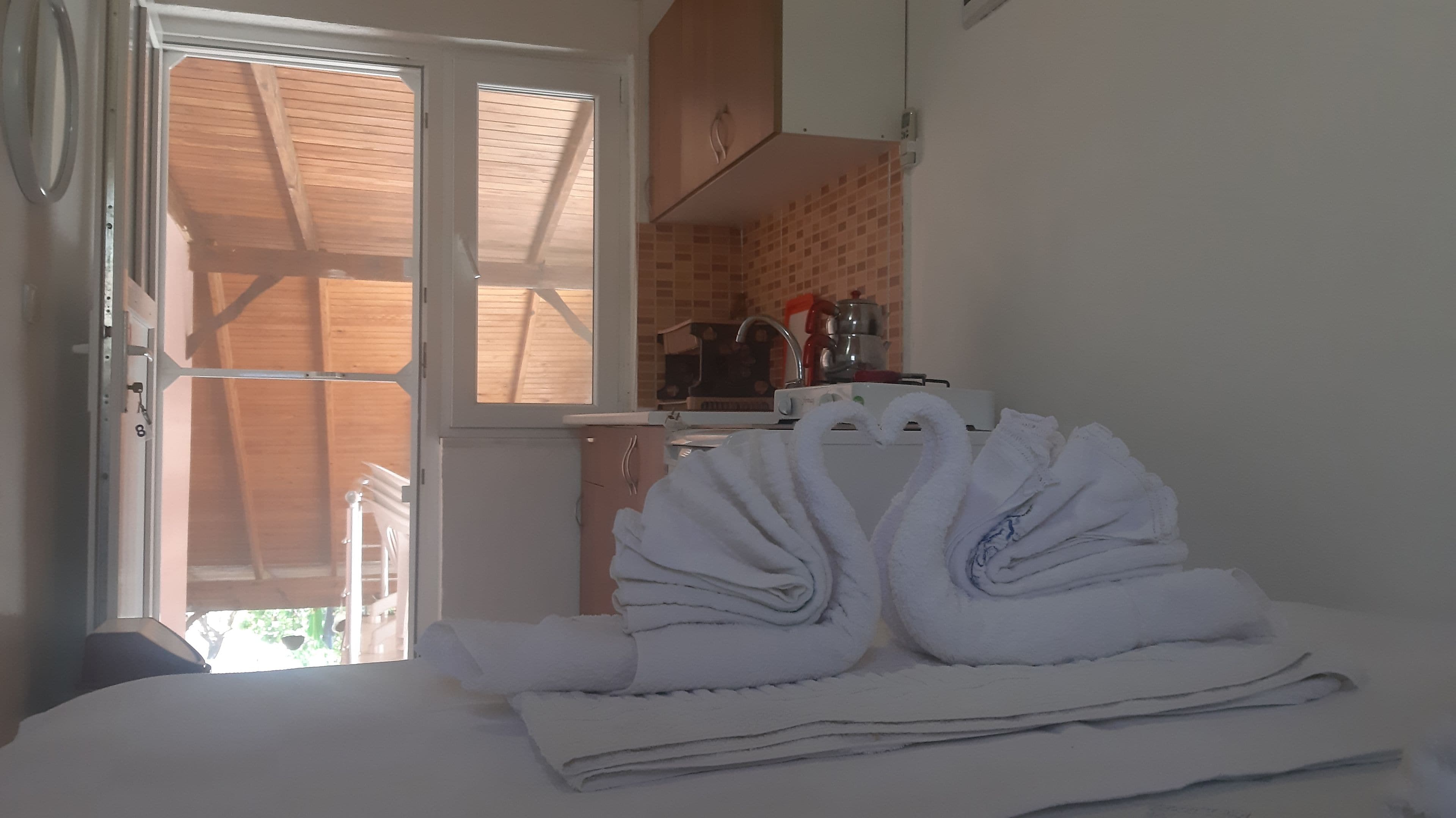 A room with a bed, a window with a view of a wooden structure outside, and a kitchenette with a stove and sink.