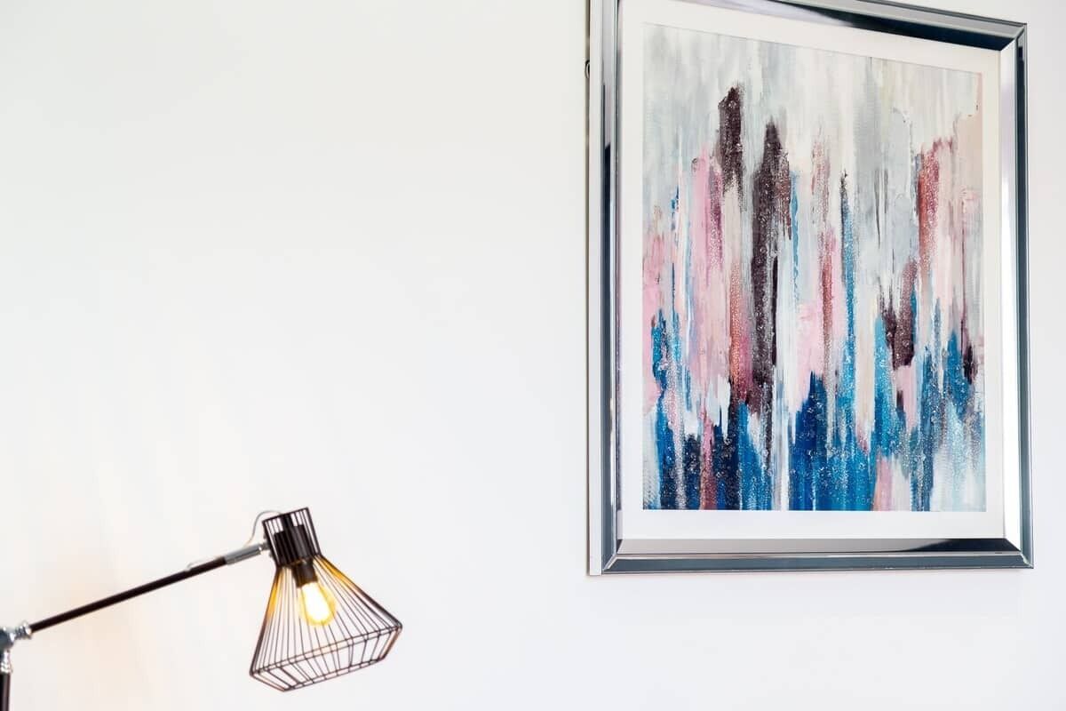 Een lijst met een abstract schilderij aan een witte muur, een zwarte bureaulamp met een glazen kap, en een lichtbulb met een metalen frame.