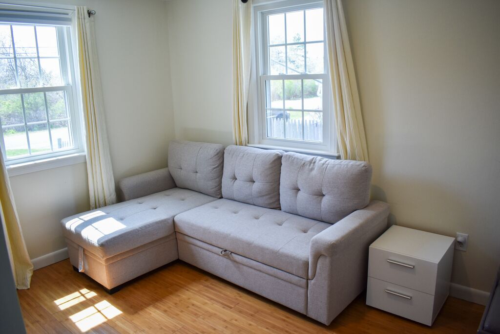Una habitación con un sofá seccional de color gris claro, una mesita de noche pequeña y una ventana con cortinas.