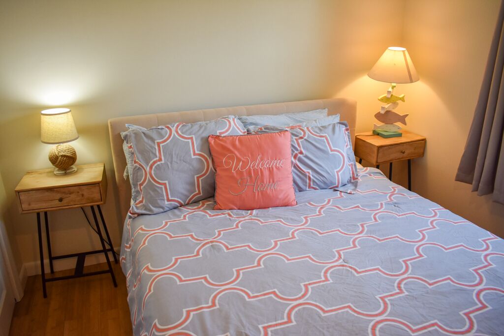 A neatly arranged bedroom with a patterned bedspread, a pillow with "Welcome Home" text, a bedside lamp, and a small wooden nightstand.