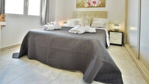 Una cama bien hecha con colcha gris, almohadas blancas y toallas dobladas sobre un suelo de baldosas.