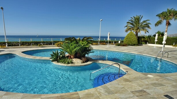 Una piscina con diseño curvo, rodeada de sillones de descanso y palmeras, con vista al mar.