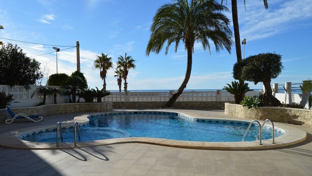 A swimming pool with a curved edge, surrounded by palm trees and a stone wall. - Alq 048 Porto Azul 2 (Calpe / Calp)