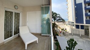 A balcony with a white lounge chair, a glass door, and a view of a building and palm trees. - Alq 048 Porto Azul 2 (Calpe / Calp)