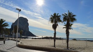 A beach with palm trees, a large rock formation, and a clear sky. - Alq 048 Porto Azul 2 (Calpe / Calp)