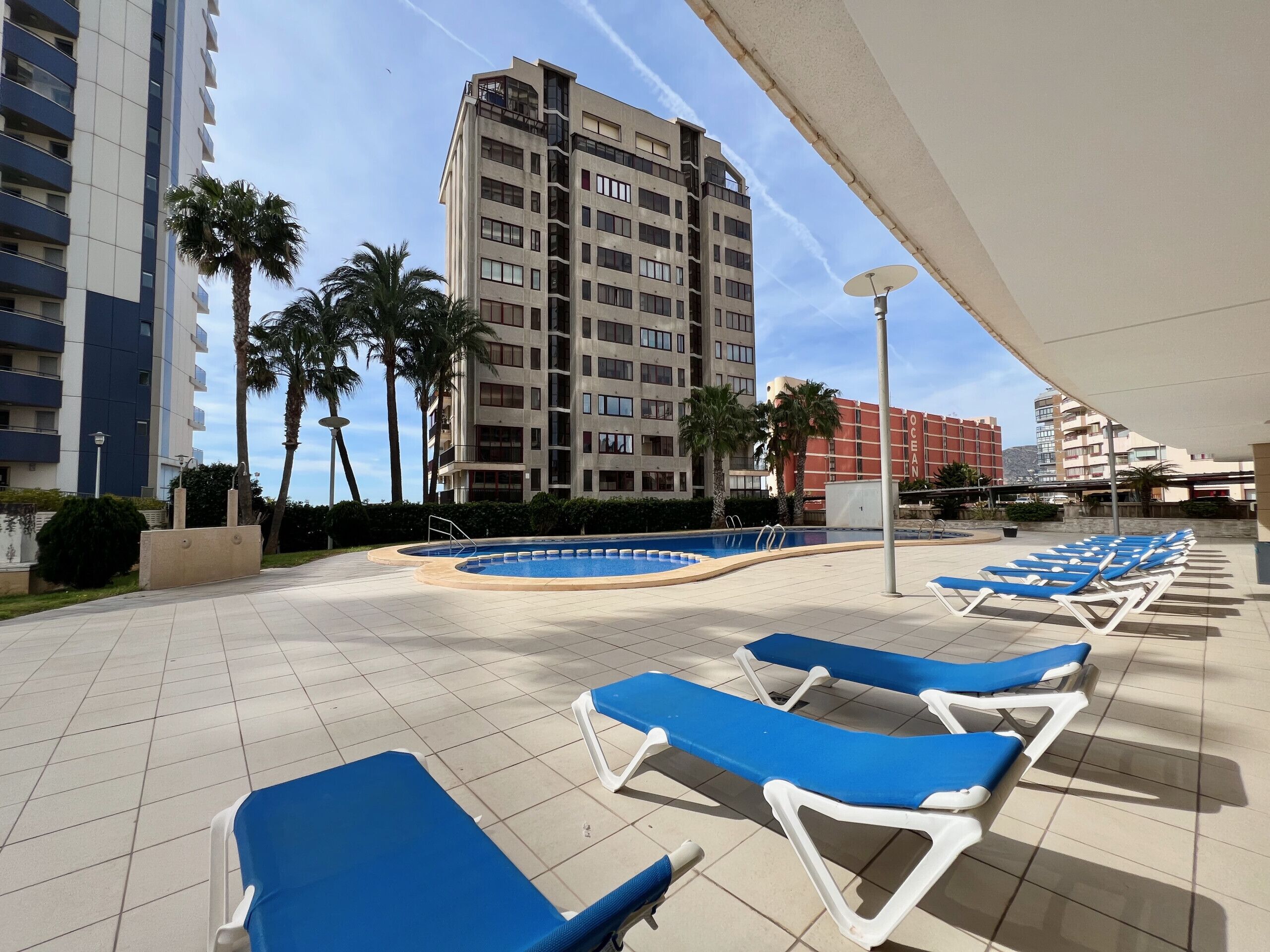 Un espace aménagé au bord de la piscine, avec des chaises longues bleues, une piscine et de grands immeubles en arrière-plan.