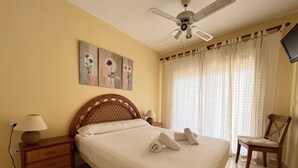A neatly made bed with pillows and towels, a wooden nightstand with a lamp, a chair, and a ceiling fan. - Alq 128 Bahia del Sol (Calpe / Calp)
