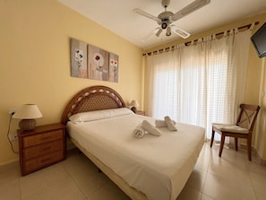 A neatly made bed with pillows and towels, a wooden nightstand with a lamp, a chair, and a ceiling fan. - Alq 128 Bahia del Sol (Calpe / Calp)