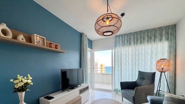 Una sala moderna con televisor, sofá y mesa de centro, un balcón con vistas, y objetos decorativos en estanterías.