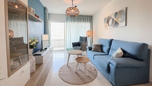 Una sala de estar moderna con un sofá azul, una mesa de centro y un televisor. Hay una ventana grande con cortinas, una lámpara de pie y un cuadro en la pared.