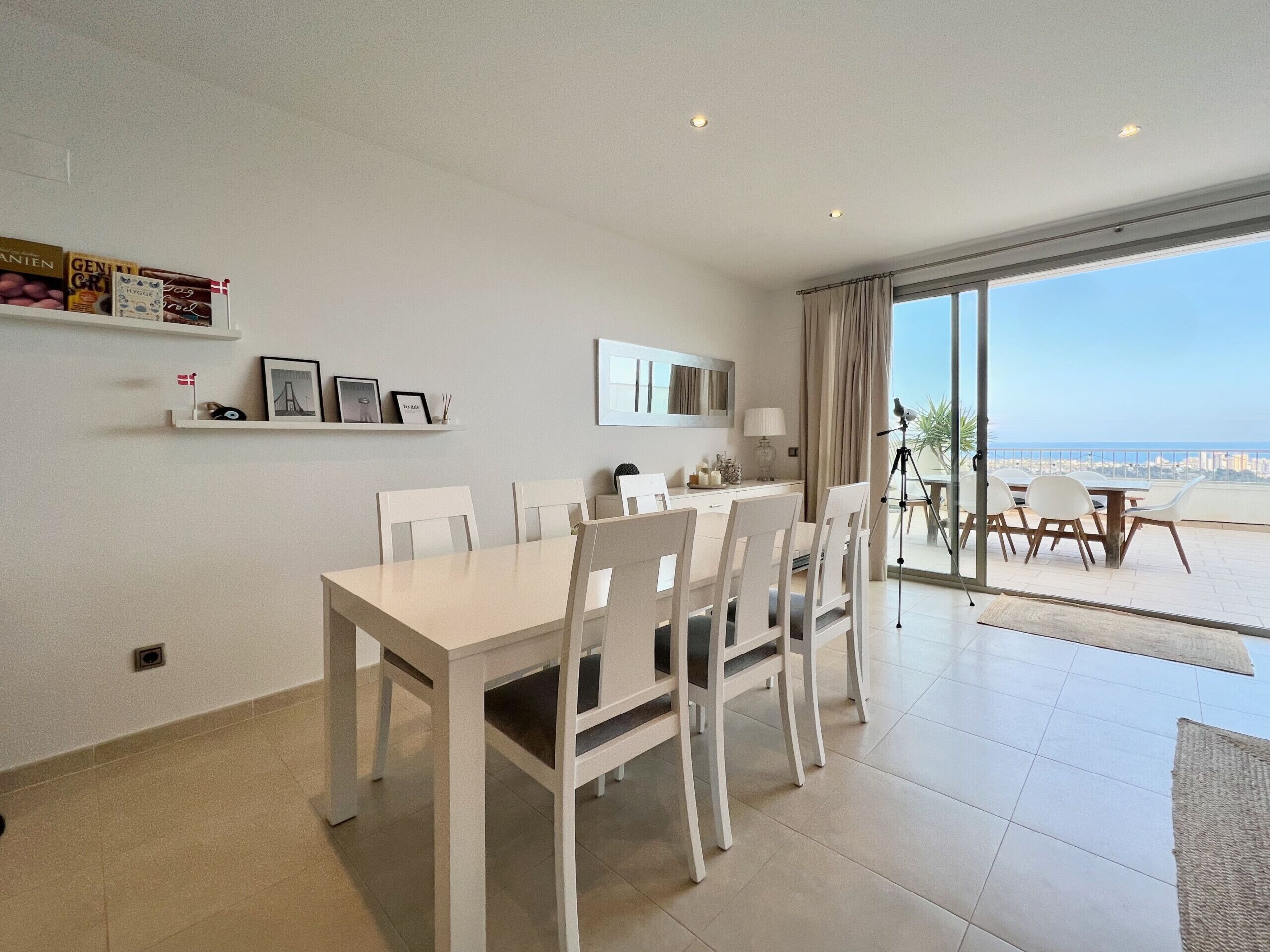 A dining area with a white table and chairs, a small shelf with items, and a view of the sea through large windows.