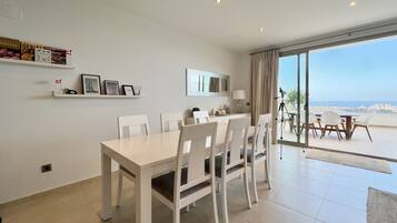 A dining area with a white table and chairs, a small shelf with items, and a view of the sea through large windows.