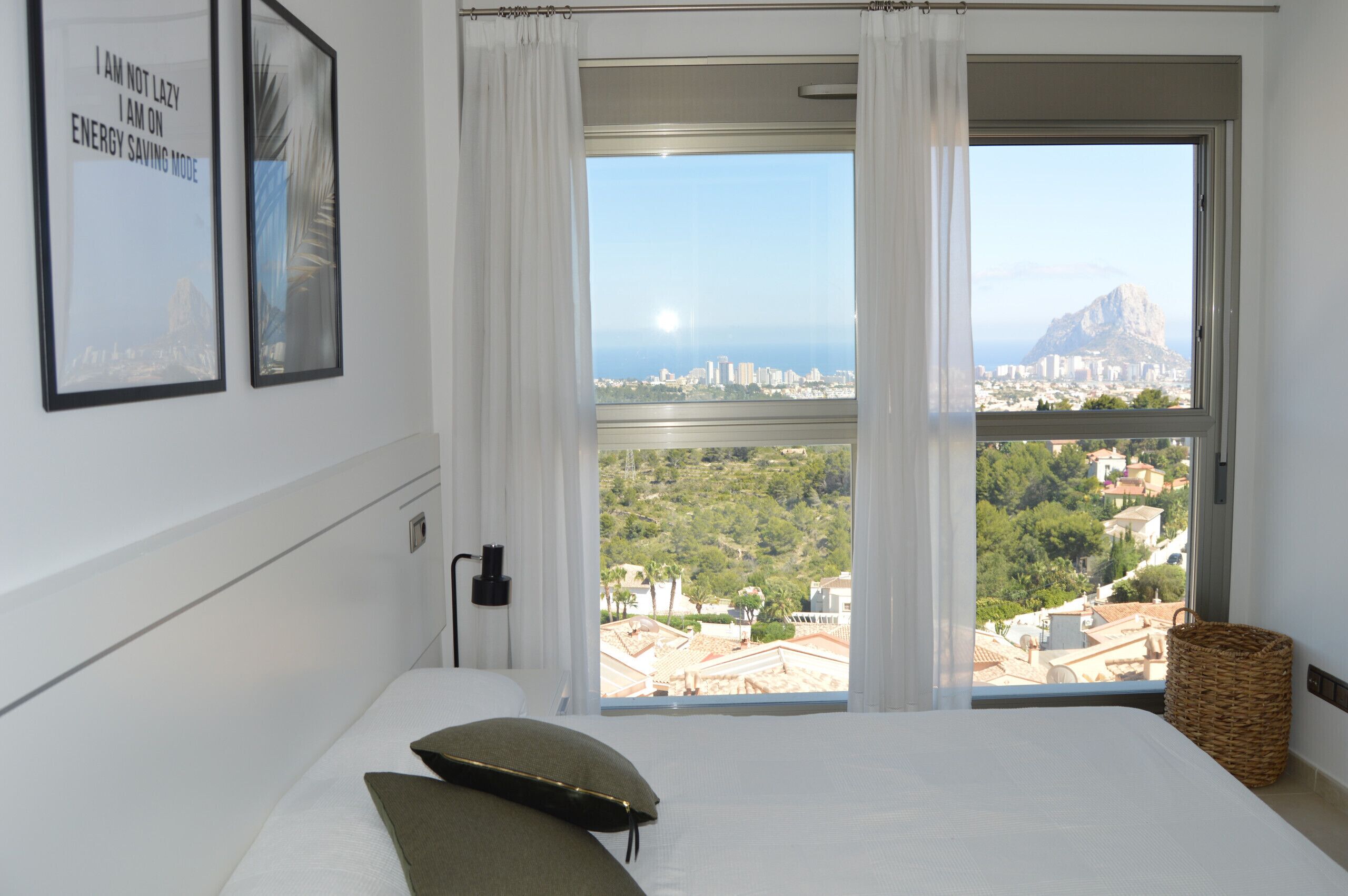 A hotel room with a large window offering a city view, a bed with pillows, and a wall-mounted power outlet.