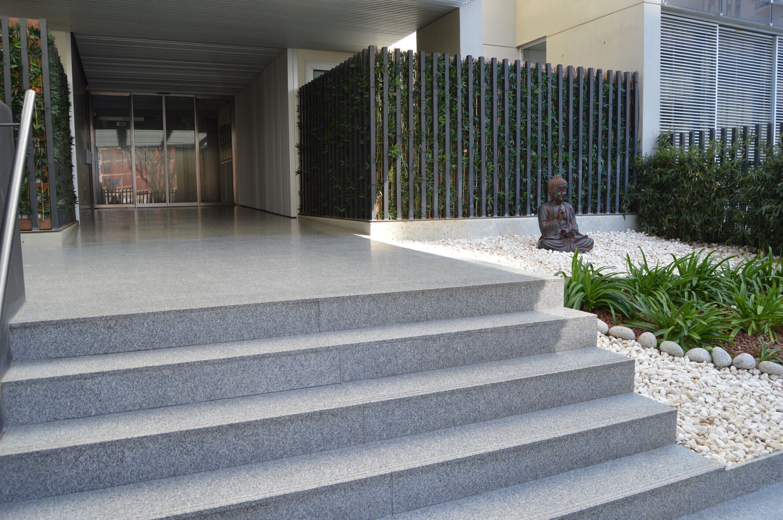 A set of stone steps leading to a building entrance, a bamboo fence, and a Buddha statue in a garden area.