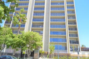 A high-rise residential building with multiple balconies, a clear sky, and surrounding greenery.