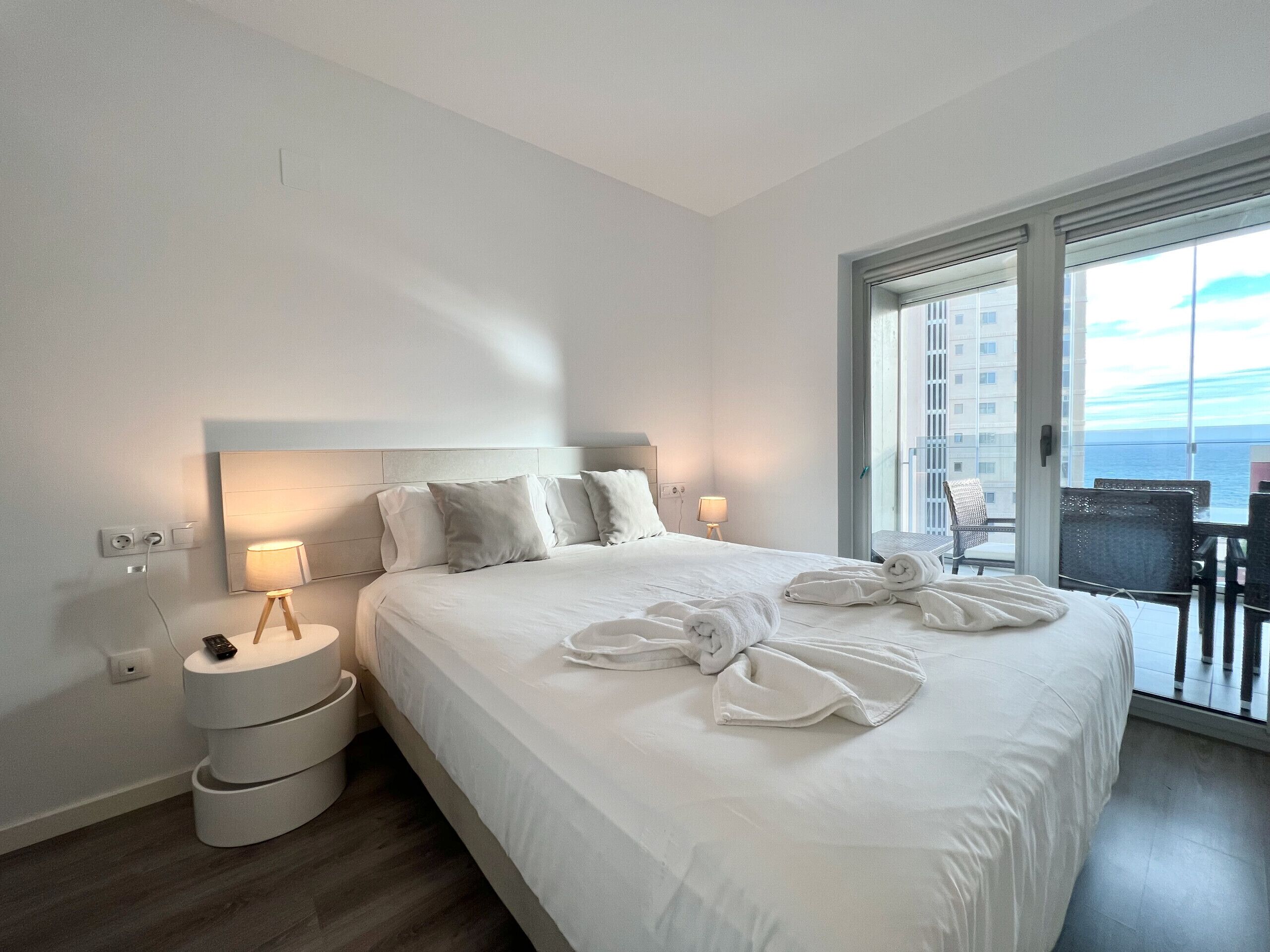 A hotel room with a large bed, a bedside table with a lamp, a power outlet, and a view of the ocean through a balcony door.