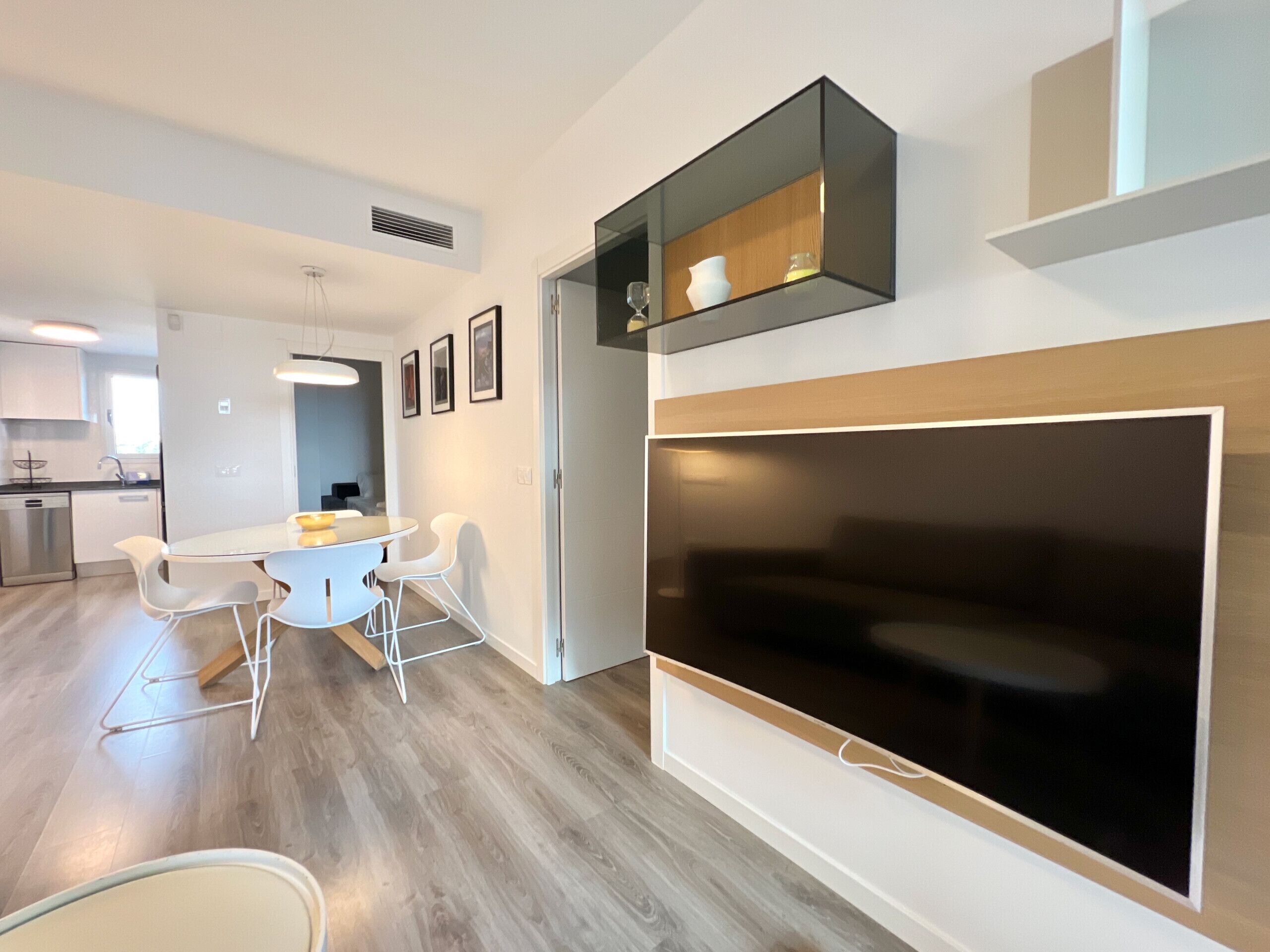 Une cuisine moderne avec des meubles blancs, un coin repas doté d’une table en verre et de chaises blanches, et un grand téléviseur à écran plat fixé au mur.