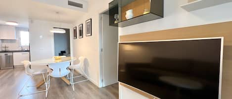 A modern kitchen with white cabinets, a dining area with a glass table and white chairs, and a large flat-screen TV mounted on the wall.