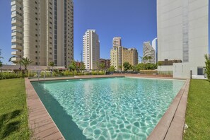 A swimming pool with a wooden deck, surrounded by grass and a fence, with tall buildings in the background.