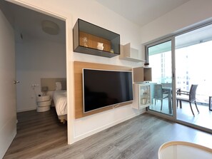 A modern hotel room with a large flat-screen TV mounted on the wall, a balcony with outdoor seating, and a wooden floor.