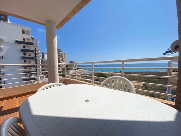 A balcony with a table and chairs, overlooking a coastal view with buildings and a clear sky.