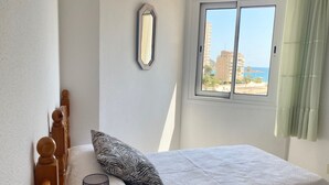 A bedroom with a bed, a wooden headboard, a patterned pillow, a wall-mounted mirror, and a window with a view of buildings and the sea. - Alq 156 Talima sea views (Calpe)