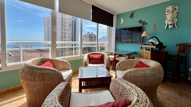 A spacious balcony with wicker furniture, a flat-screen TV, and a view of buildings and the sea.