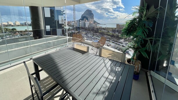 En balkon med et træbord og stole, en potteplante og udsigt over et bybillede med bygninger og et bjerg.