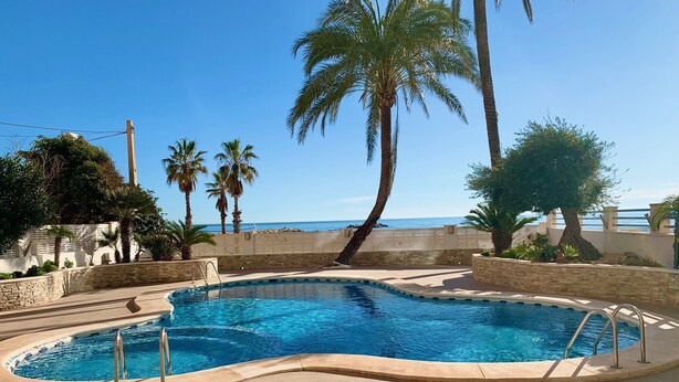 A swimming pool with a curved edge, surrounded by stone walls and palm trees. - Alq 043 Porto Azul 4 (Calpe)