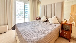 A modern hotel bedroom with a large bed, a wooden nightstand, a mirror, and a view of the city through the window. - Alq 043 Porto Azul 4 (Calpe)