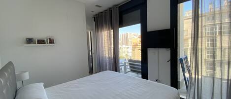 A modern hotel room with a large bed, a balcony with a view of the city, and a door leading to an outdoor area.