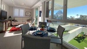 A modern living room with a glass dining table set for four, a sofa, a TV, and a balcony with a view of the city.