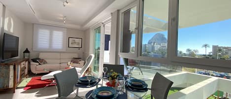 Ein modernes Wohnzimmer mit einem Glastisch, gedeckt für vier Personen, einer Couch, einem Fernseher und einem Balkon mit Blick auf die Stadt.