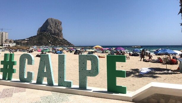A beach with a large rock formation in the background, a sign with '#CALPE', and people relaxing under umbrellas.